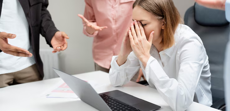 Employee holding her head while a colleague bullies and criticizes her verbally, illustrating how verbal threats can escalate to violence.